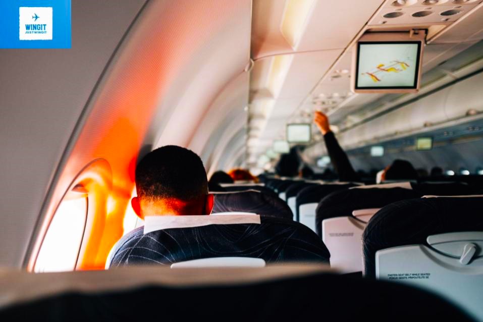 The back of people's heads as they wait for takeoff on a commercial airplane. One person has their hand raised like they are asking as question.